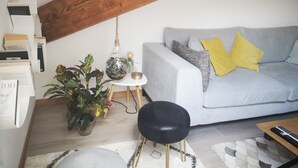 A living room with a grey sofa, yellow cushions, a black chair, a wooden ceiling beam, and a hanging plant.