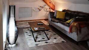 A cozy loft bedroom with a sloped ceiling, a wooden beam, a small window, a sofa with cushions, a floor lamp, and a coffee table.