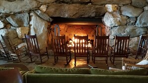 Une cheminée en pierre avec un feu, entourée de chaises en bois et d’un canapé vert.