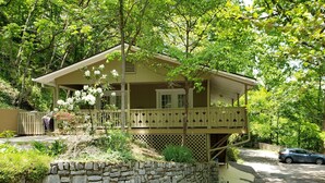 Une maison à deux étages dotée d’un balcon, entourée d’une végétation luxuriante et d’un mur de pierre.