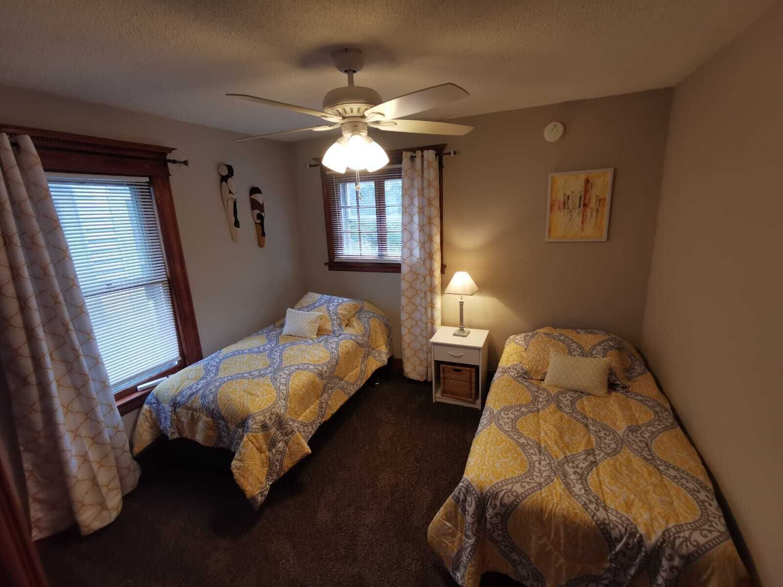 Un dormitorio con dos camas individuales, un ventilador de techo, una mesita de noche con lámpara y un cuadro en la pared.