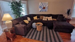 A living room with a dark brown sectional sofa, a glass coffee table, a striped rug, a ceiling fan, and a large window with blinds.