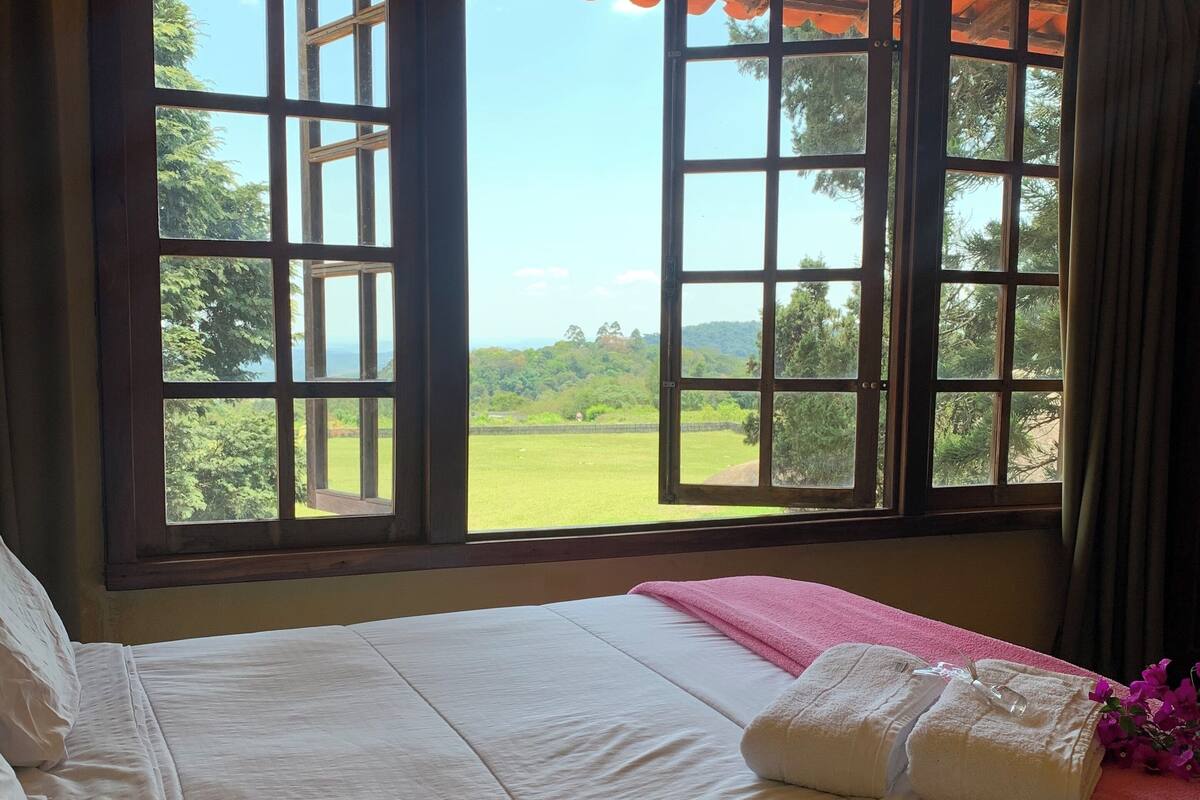 A bedroom with a large window, a bed with white linens, and a pink blanket.