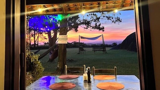 A framed view of a sunset scene with a hammock and a table with bottles, under a thatched roof structure. - Pousada Refúgio Dinamarca (Mairipora)