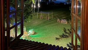 A view from a wooden-framed window overlooking a well-lit outdoor area with a grassy lawn, a hammock, and a small table. - Pousada Refúgio Dinamarca (Mairipora)