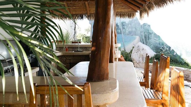 A wooden outdoor seating area with a thatched roof, stone table, and chairs.