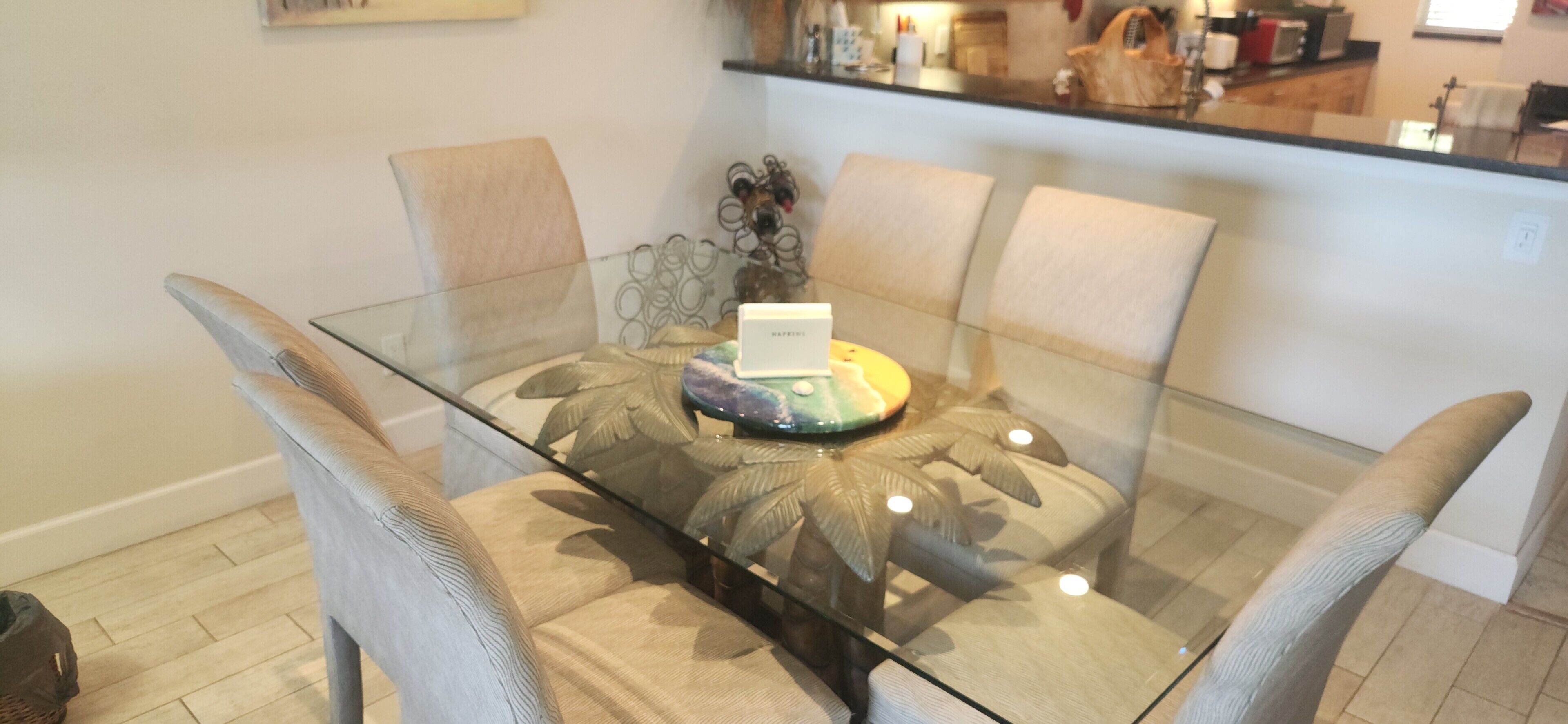 Un coin repas avec une table en verre entourée de chaises rembourrées, un élément décoratif au centre et un espace cuisine en arrière-plan.