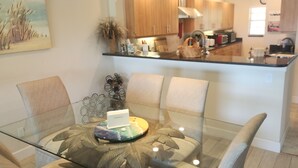 Un comedor con mesa de vidrio, sillas acolchadas, un centro de mesa decorativo y una cocina al fondo.