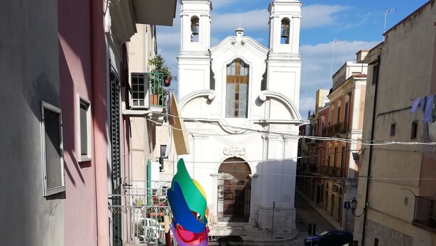 Une rue étroite, avec une église blanche en arrière-plan, une voiture bleue garée sur le côté et un trottoir pour les piétons.