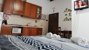 Une chambre à coucher comprenant un lit, un téléviseur, des armoires en bois, un coin cuisine et une porte ornée d’une croix.