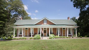A two-story brick house with a green roof, a front porch with white columns, and a flag.