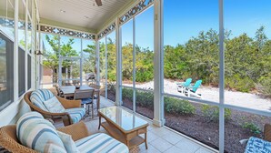 A covered patio with wicker furniture, a glass table, and a view of a sandy area with greenery. - Bella Vue~ Stunning 3BR/3 1/2 BA beach house! (Gulf Shores)
