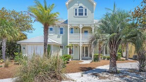 A two-story house with a porch, surrounded by palm trees and a sandy driveway. - Bella Vue~ Stunning 3BR/3 1/2 BA beach house! (Gulf Shores)
