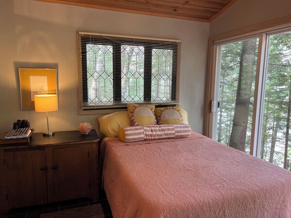 Une chambre à coucher comprenant un lit, une table de chevet, une lampe, une fenêtre avec des stores et une vue sur les arbres.