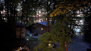 Een rustieke hut met een zandpad er naartoe, omringd door dichte bomen en een kleine vijver.