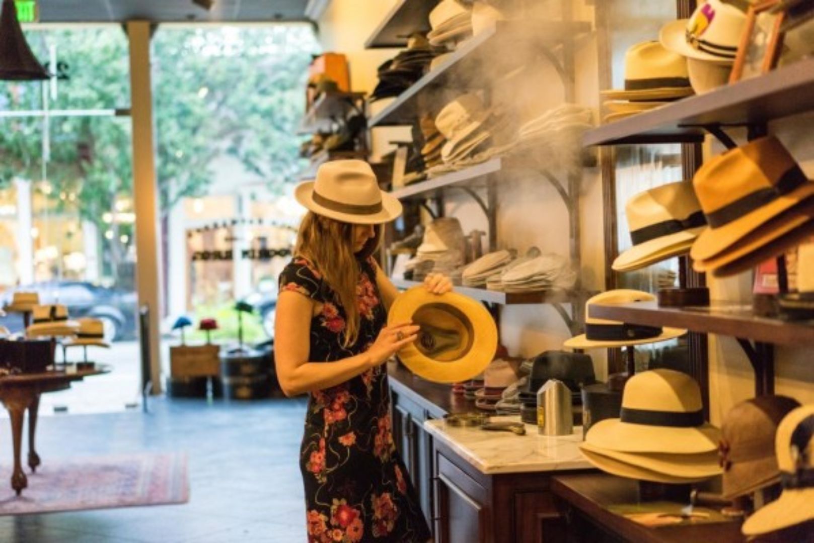 A person examining hats in a store with shelves of hats and a view of the outside.