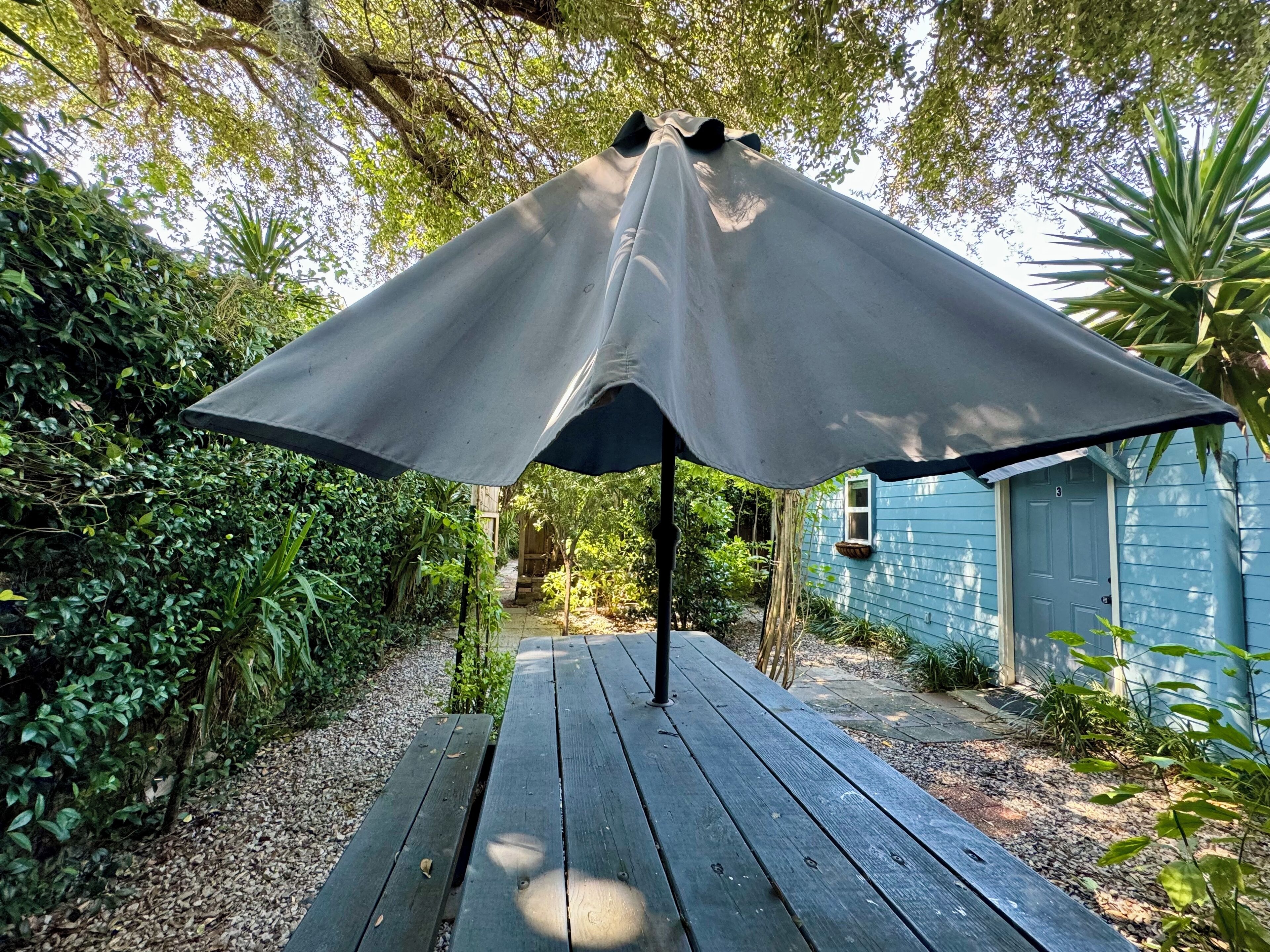 Una terraza de madera con una amplia estructura tipo sombrilla, rodeada de exuberante vegetación y una casa azul al fondo.