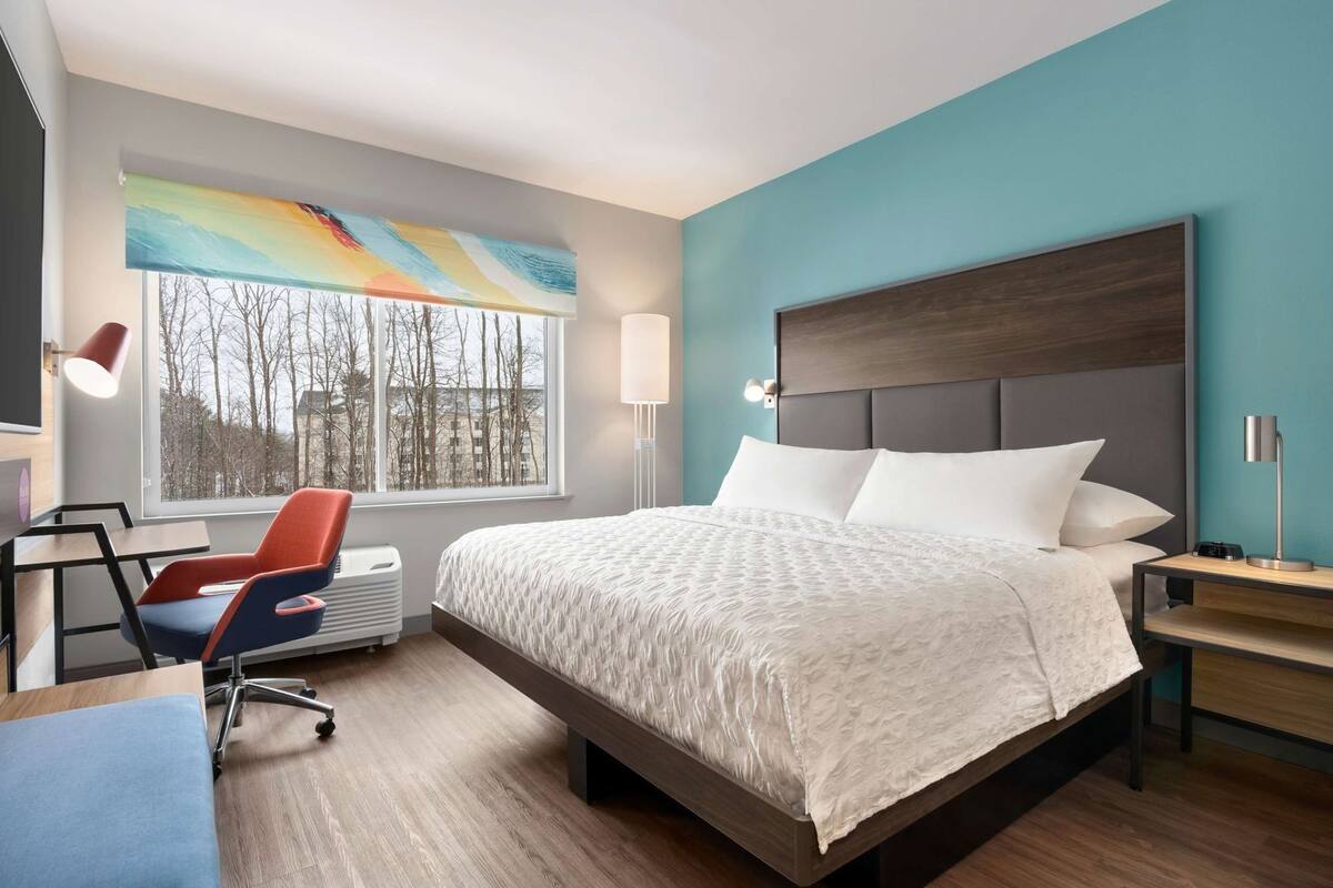 Une chambre d’hôtel moderne avec un grand lit, un bureau avec une chaise, une fenêtre donnant sur des arbres et une œuvre d’art colorée accrochée au mur.
