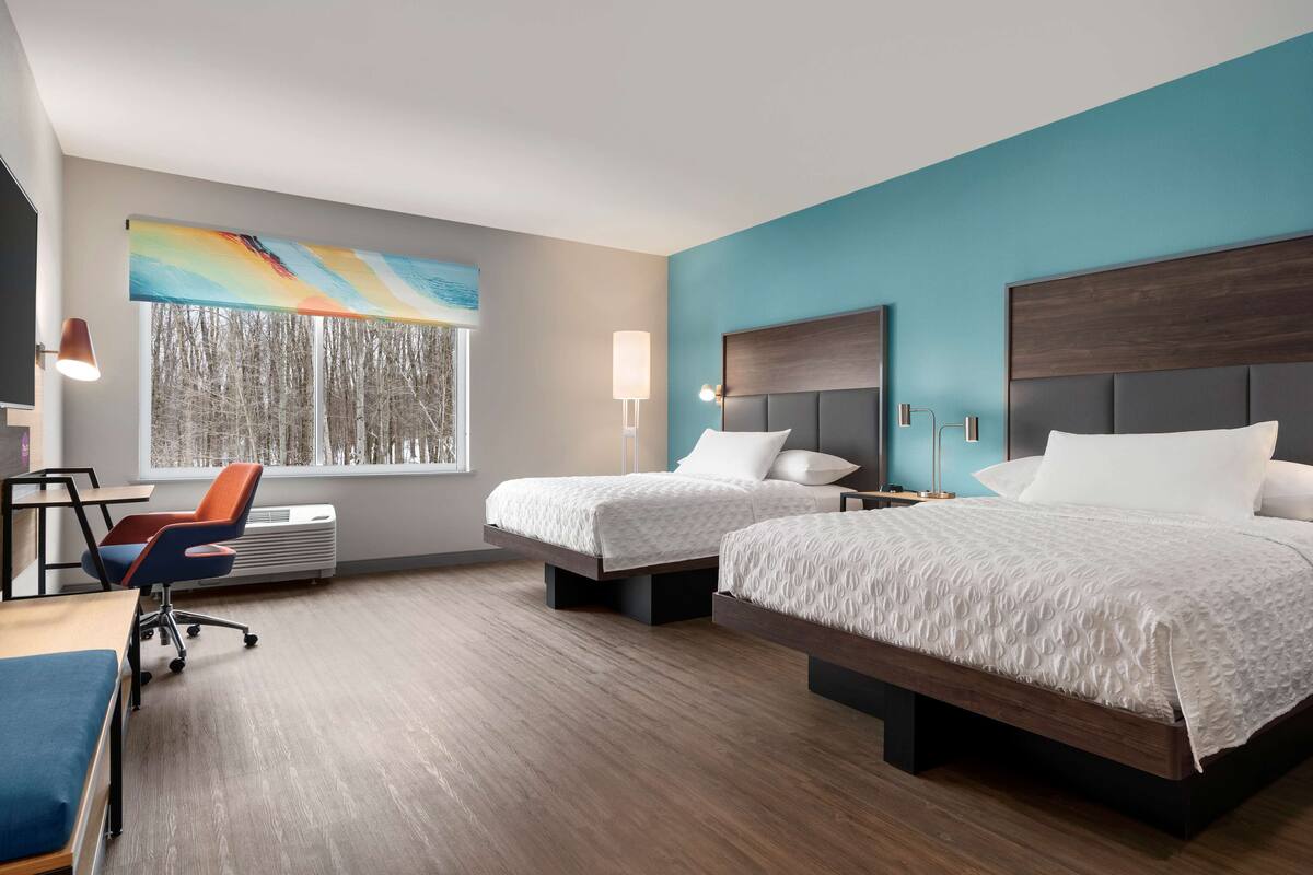Een hotelkamer met twee bedden, een bureau, een stoel en een groot raam met uitzicht op bomen.