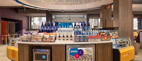 Una estación autoservicio de bebidas con una sección refrigerada, diversos aperitivos y un terminal de pago digital.