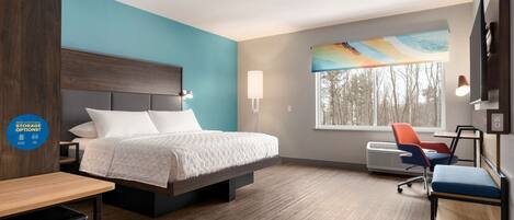 A hotel room with a bed, a desk, a chair, a TV, and a window with a view of trees.