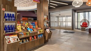 Un espacio comercial moderno con estanterías de madera repletas de aperitivos y bebidas, un letrero de mercado abierto las 24 horas, y una zona de recepción con puerta de vidrio de acceso.