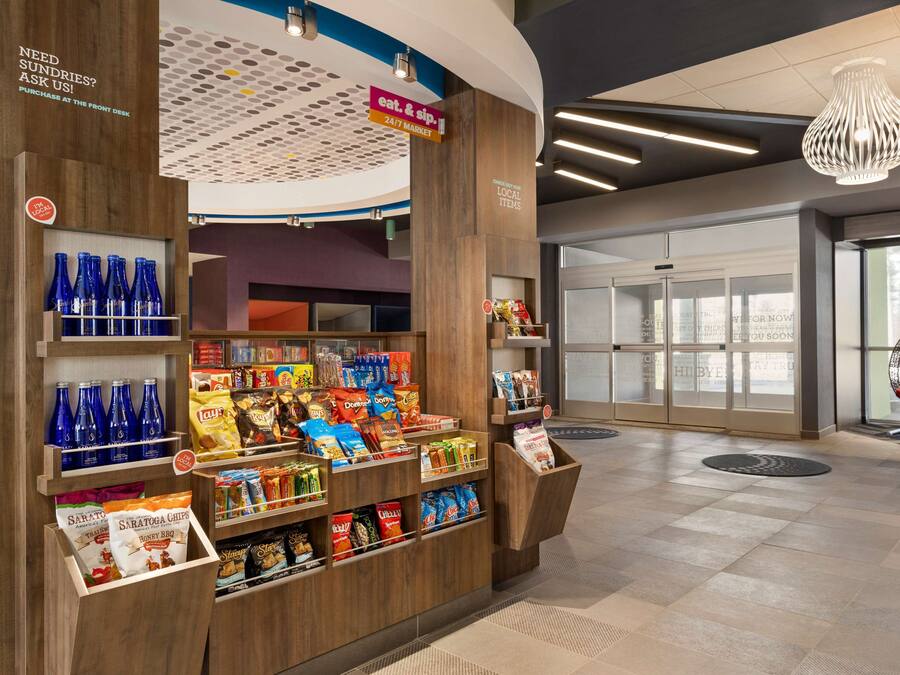 A modern retail area with wooden shelving stocked with snacks and beverages, a 24/7 market sign, and a front desk area with a glass door entrance.