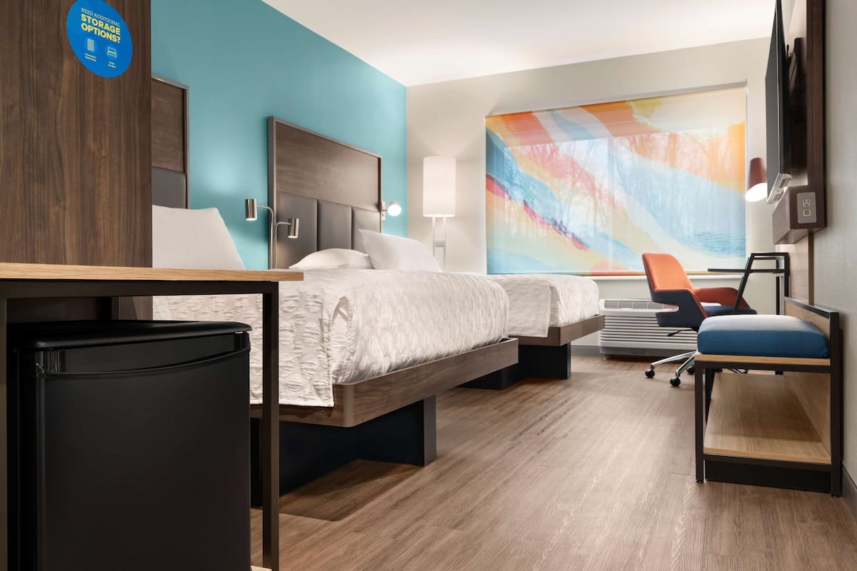 Een hotelkamer met twee bedden, een bureau, een stoel, een koelkast, en een groot abstract schilderij aan de muur.
