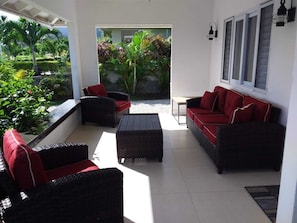 Un espacio al aire libre con sol, mobiliario de mimbre y cojines rojos, con vistas a un jardín exuberante.