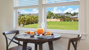 A breakfast table with a pitcher of orange juice, a bowl of fruit, and two glasses of juice, set against a window with blinds.