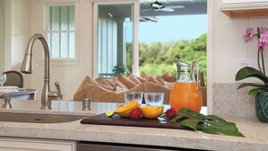 A kitchen with a stainless steel faucet, a wooden cutting board with fruit, a pitcher of orange juice, and a view of a balcony with a ceiling fan. - Hale Lo'i + Cottage: 5BR home +pool in Sunny Poipu (Koloa)