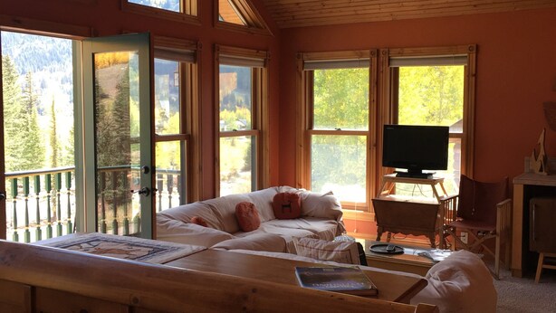 A bedroom with a large bed, a TV, and a desk. There are multiple windows providing a view of the outdoors.
