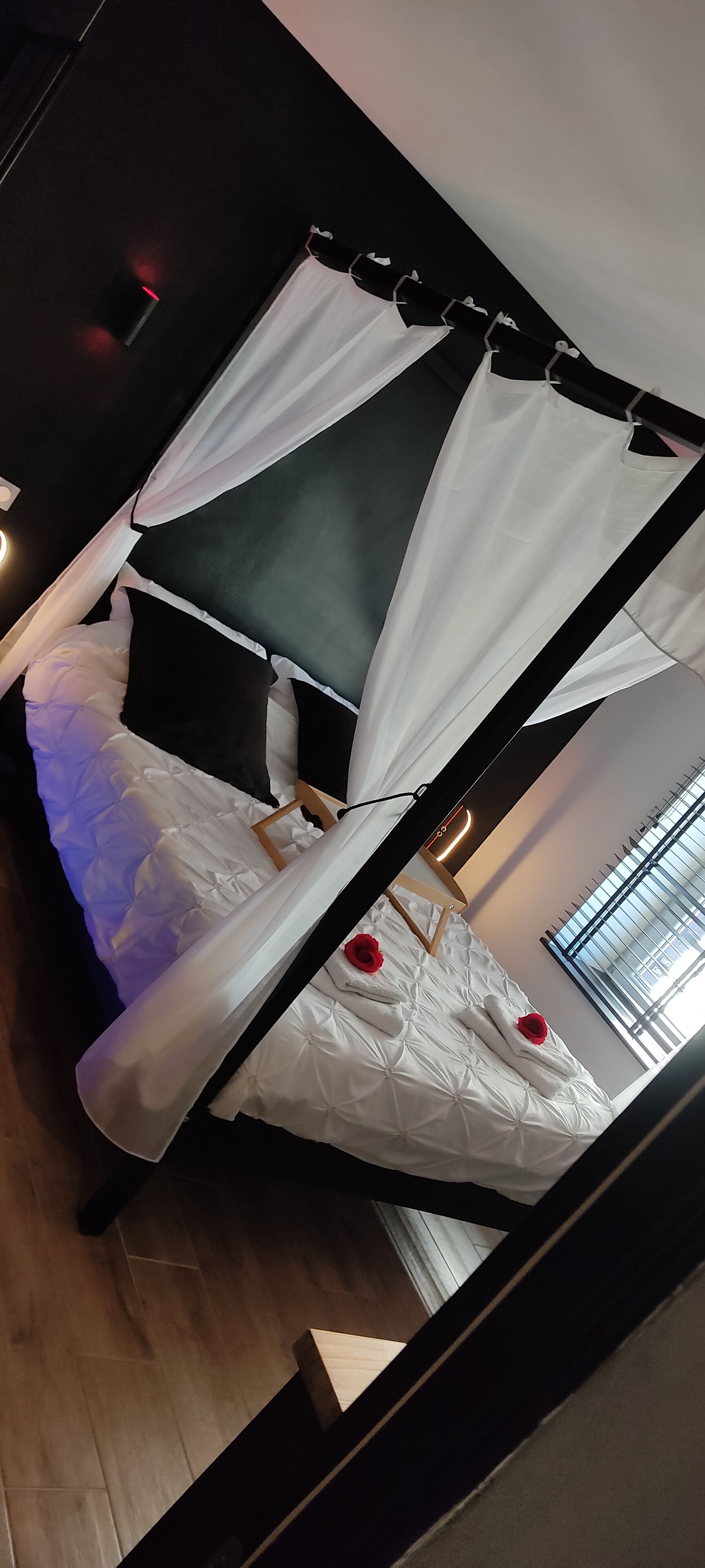 Ein Himmelsbett mit weißer Bettwäsche und schwarzen Kissen, ein Nachttisch mit Lampe sowie ein Fenster mit Jalousien.