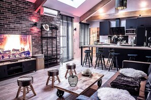 Una cocina moderna con armarios negros, techo de madera y un televisor grande. Hay taburetes y una mesa con un florero.
