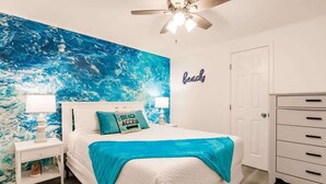 A bedroom with a large ocean-themed mural, a bed with a blue throw, a nightstand with a lamp, and a dresser. - Coconut Dreams! Heated Pool, 4 min to AMI Beach (Bradenton)