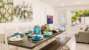 A dining area with a long table set for a meal, white walls, and a large window with a view of palm trees. - Coconut Dreams! Heated Pool, 4 min to AMI Beach (Bradenton)