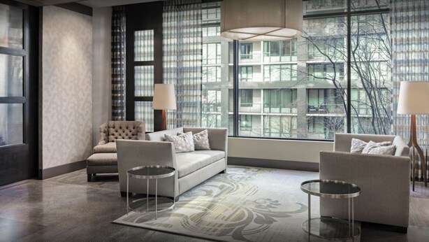 Una sala de estar moderna con un sofá, sillones y una mesa de centro. Hay ventanas amplias que ofrecen vistas a edificios y árboles.