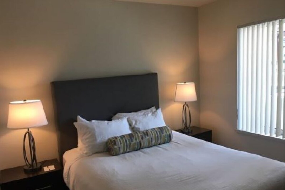 Cama bem arrumada com lençóis brancos e duas almofadas, com duas mesas de cabeceira com abajures, em um quarto com janela e paredes lisas.
