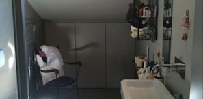 Un bagno piccolo con doccia, lavandino e specchio. Nell'angolo c'è una sedia e uno scaffale con articoli da toilette.