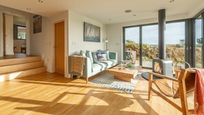 A spacious living room with a wooden floor, a sofa with cushions, a coffee table, and a large window with a view of the outdoors. - A stunning two bedroom eco-home with breath-taking countryside views. (Chapel Porth)