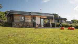 A modern house with a stone facade, a wooden deck, and a grassy yard with yellow flowers. - A stunning two bedroom eco-home with breath-taking countryside views. (Chapel Porth)