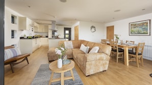 Un salon moderne avec un canapé d’angle beige, une table à manger en bois avec des chaises, une cuisine en arrière-plan et un tableau accroché au mur.