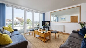 A modern living room with a sofa, coffee table, and TV, large windows, and a view of houses. - A superbly situated property that is just a short walk from two fabulous beaches (Newquay)
