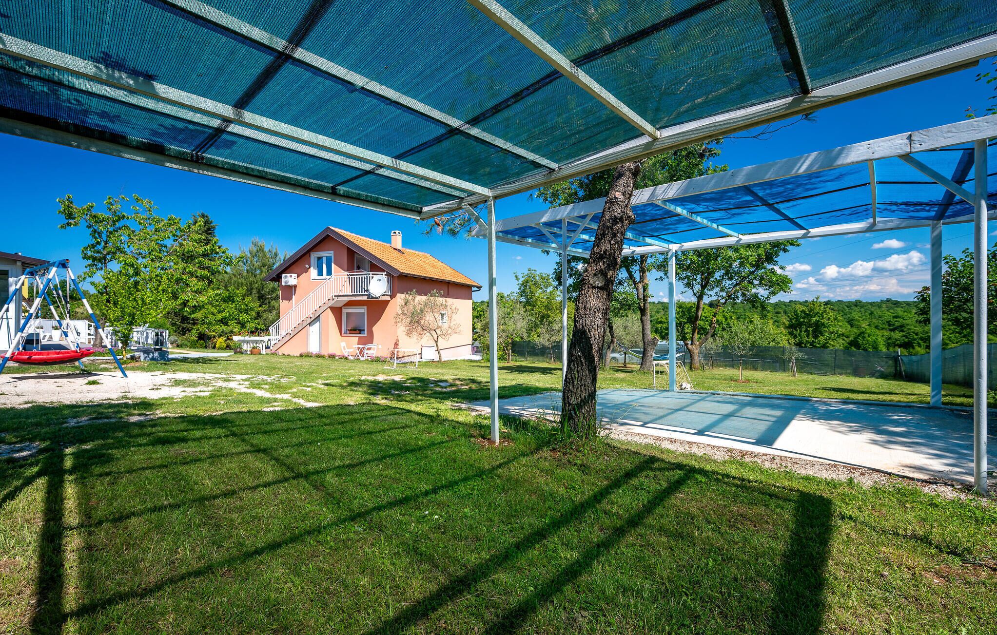 A covered outdoor area with a house in the background, a tree, and a swing set.