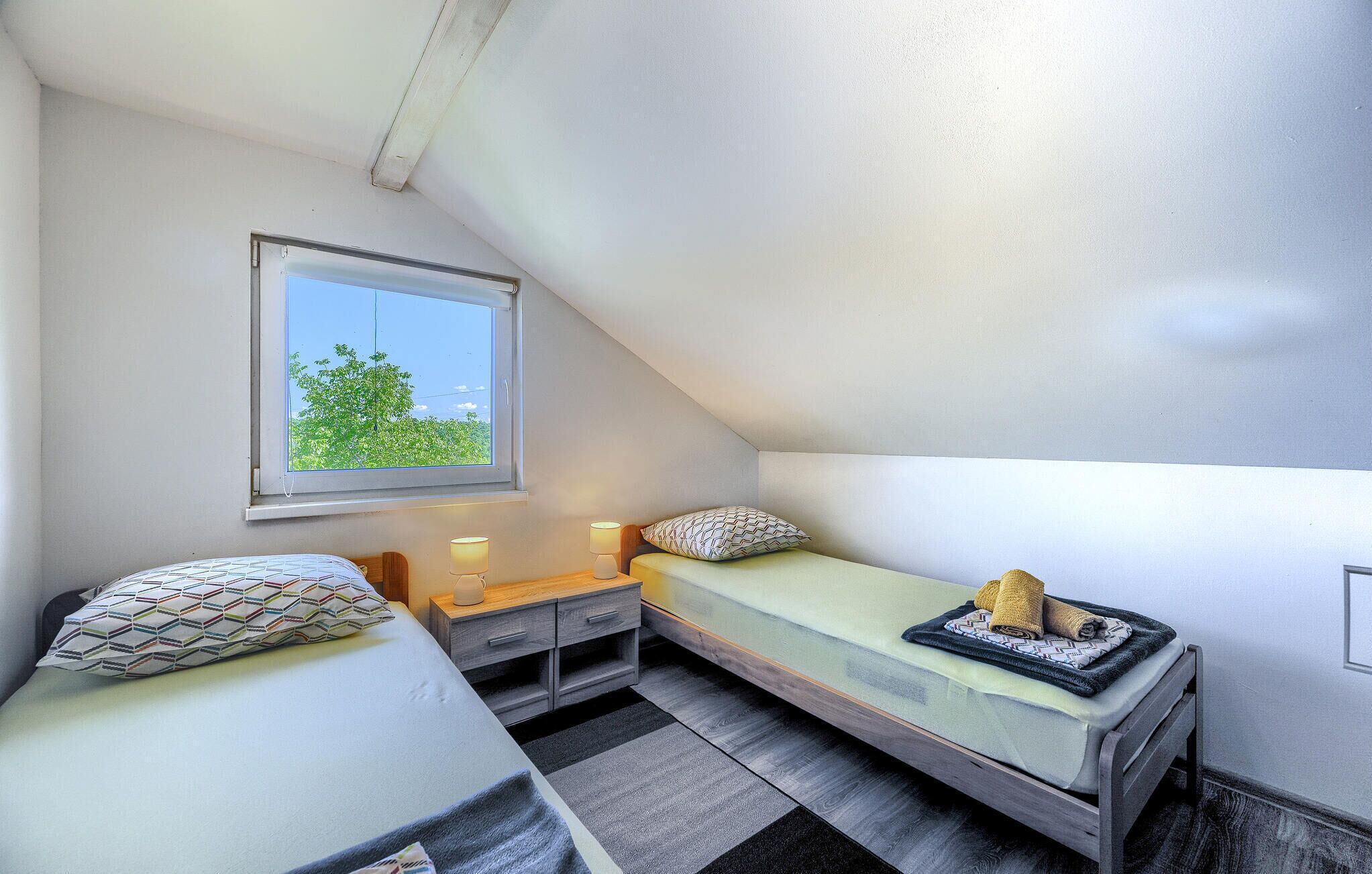 Een kleine, compacte kamer met een schuine kap, een enkel venster met uitzicht op groen, een bed met een gekleurd kussen, een nachtkastje met een lamp, en een klein nachtkastje met een handdoek.