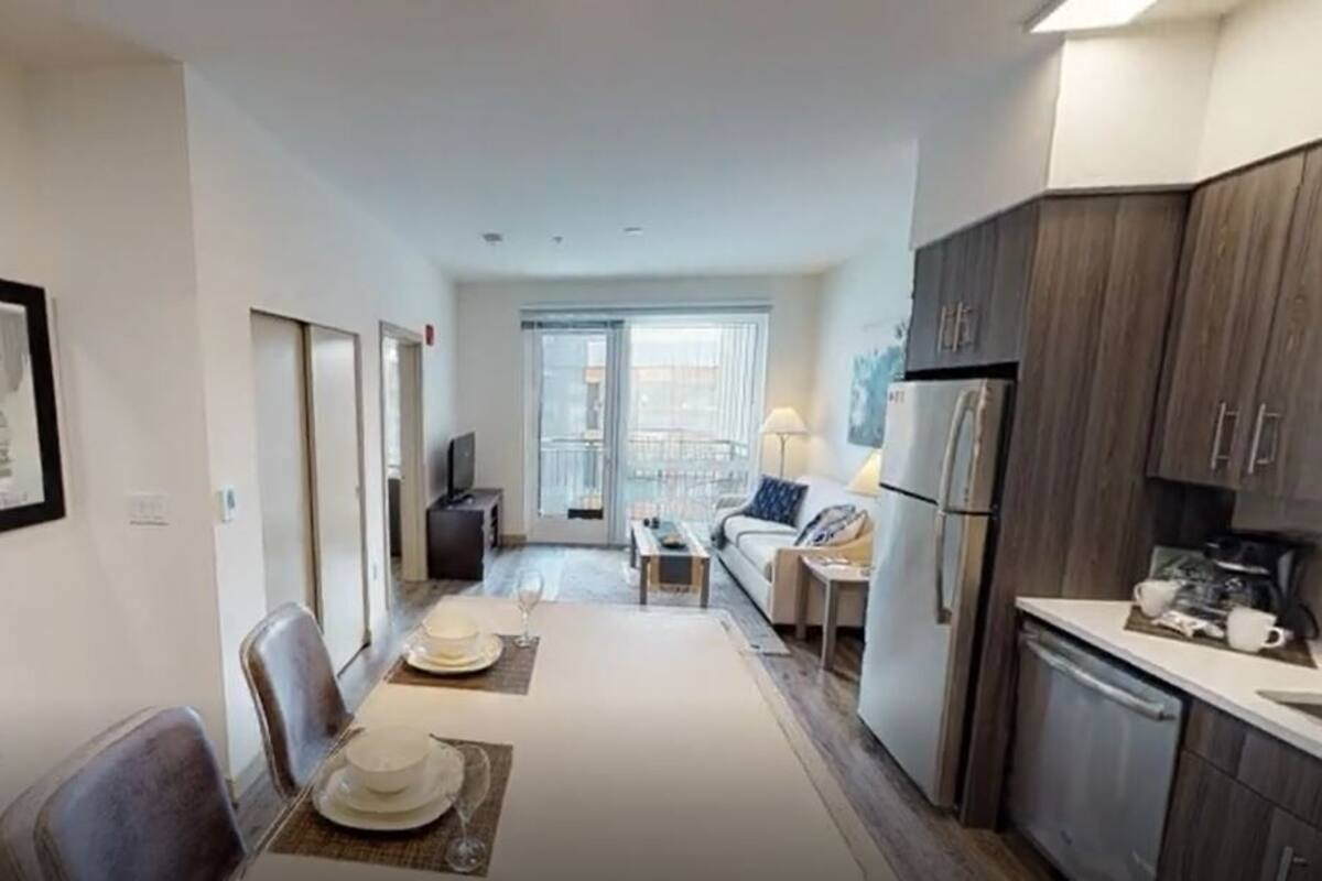 A compact hotel room with a kitchenette, a dining area with a table set for two, and a small seating area with a sofa and a TV.