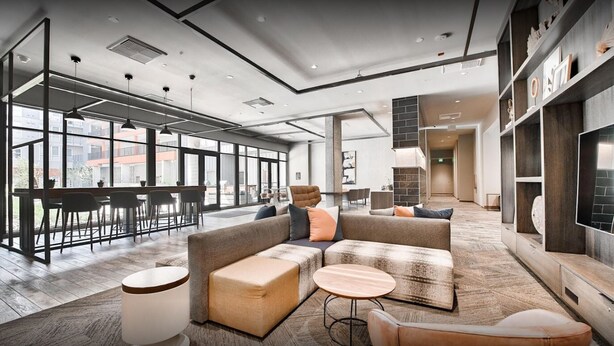 A spacious lounge area with a large sofa, ottomans, and a round table. There are multiple seating arrangements and a view of the outside through large windows. - Bell Overlake (Redmond)