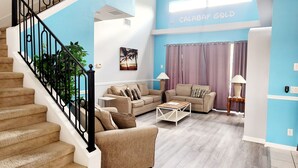 Un salon avec un escalier, des canapés de couleur beige, une table basse et un ventilateur de plafond.