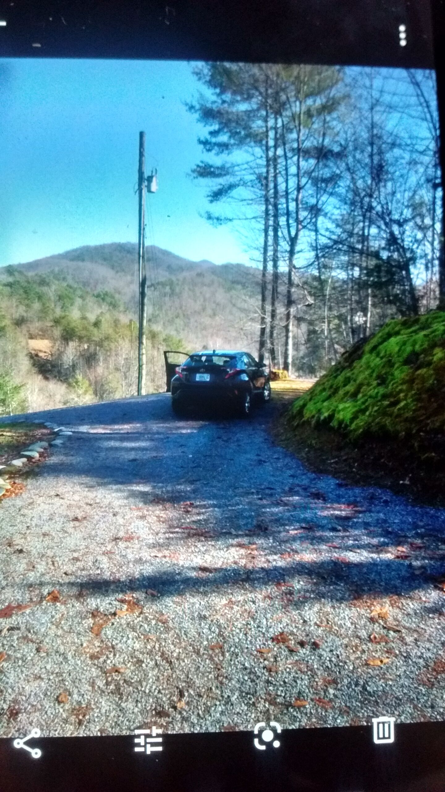 Ein Auto parkt auf einer Schotterstraße, im Hintergrund ein Wald und Berge.