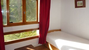 Une petite chambre avec un lit simple, un banc en bois, de grandes fenêtres et un tableau encadré au mur.